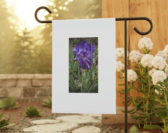 May include: A white garden flag featuring a vibrant image of a purple iris flower with white streaks. The flag is displayed on a black metal stand in a garden setting, surrounded by greenery and stone pathways. The iris is in full bloom.