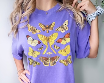May include: A lavender-colored t-shirt featuring a collection of illustrated butterflies in various sizes and colors, including yellow, brown, and orange. The butterflies are arranged across the front of the shirt, creating a nature-inspired design.