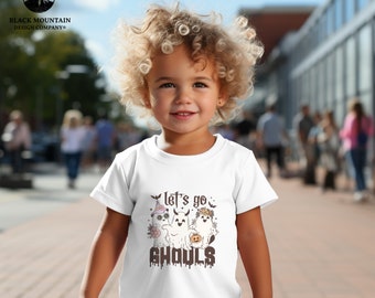 Può includere: T-shirt bianco per bambini piccoli con una grafica di tre fantasmi dei cartoni animati e la scritta "Let's go Ghouls". I fantasmi sono decorati con cappelli da strega, fiori e una zucca. La maglietta ha maniche corte.