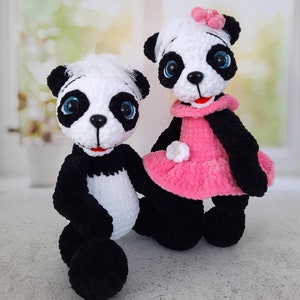 May include: Two crocheted panda plushies, one wearing a pink dress with white flowers and a blue flower on its head, the other with black and white fur.