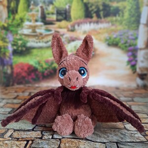 Pu&ograve; includere: Un giocattolo di peluche a forma di pipistrello in crochet marrone con grandi occhi blu e una bocca rosa. Il giocattolo ha una pelliccia marrone e grandi ali.