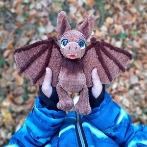 Pu&ograve; includere: Un pipistrello di uncinetto marrone con grandi occhi blu e un viso peloso marrone. Il pipistrello &egrave; tenuto da una persona che indossa una giacca blu.