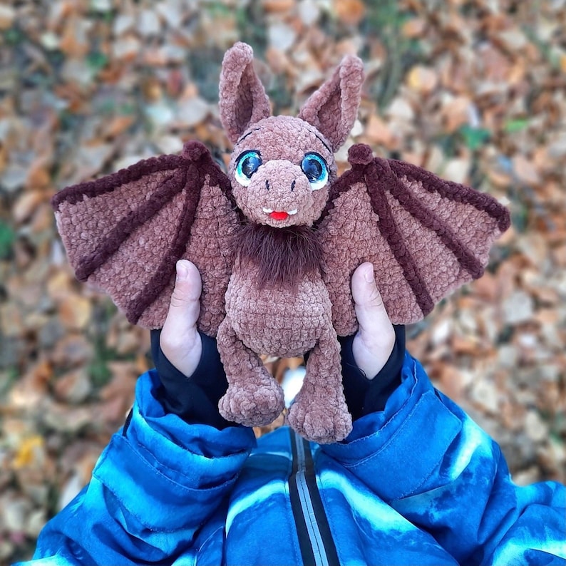 Pu&ograve; includere: Un pipistrello di uncinetto marrone con grandi occhi blu e un viso peloso marrone. Il pipistrello &egrave; tenuto da una persona che indossa una giacca blu.