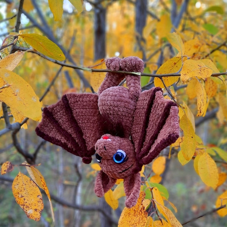 Pu&ograve; includere: Un pipistrello di uncinetto marrone con grandi occhi blu appeso a un ramo. Il pipistrello ha una consistenza pelosa ed &egrave; un'artigianato fatto a mano.