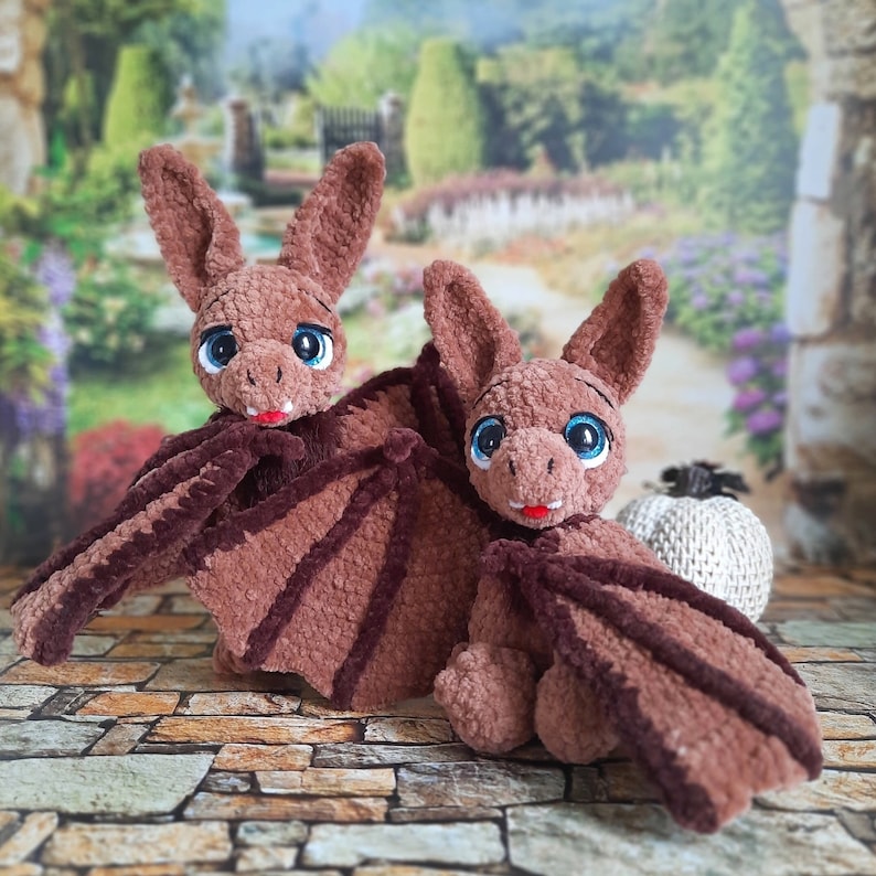 Pu&ograve; includere: Due peluche di pipistrello in crochet marrone con grandi occhi blu e nasi rosa. I pipistrelli sono seduti su una superficie di pietra.