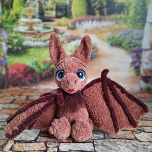 Pu&ograve; includere: Un giocattolo di peluche a forma di pipistrello in crochet marrone con grandi occhi blu e una lingua rosa. Il giocattolo &egrave; seduto su una superficie di pietra con uno sfondo di giardino sfocato.