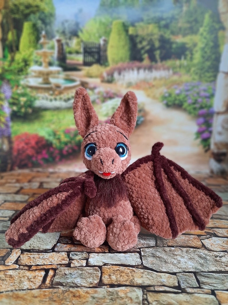 Pu&ograve; includere: Un giocattolo di peluche a forma di pipistrello in crochet marrone con grandi occhi blu e una lingua rosa. Il giocattolo &egrave; seduto su una superficie di pietra con uno sfondo di giardino sfocato.