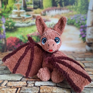 Pu&ograve; includere: Un giocattolo di peluche a forma di pipistrello in crochet marrone con grandi occhi blu e una bocca rossa. Il giocattolo &egrave; seduto su una superficie di pietra.