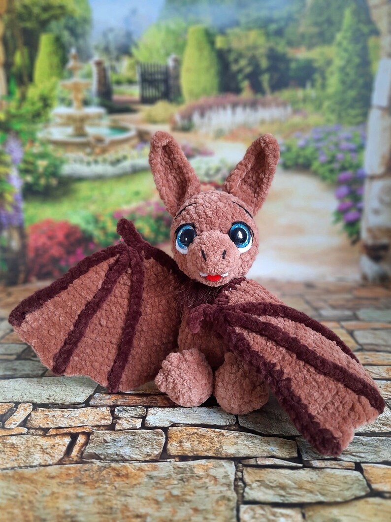 Pu&ograve; includere: Un giocattolo di peluche a forma di pipistrello in crochet marrone con grandi occhi blu e una bocca rossa. Il giocattolo &egrave; seduto su una superficie di pietra.