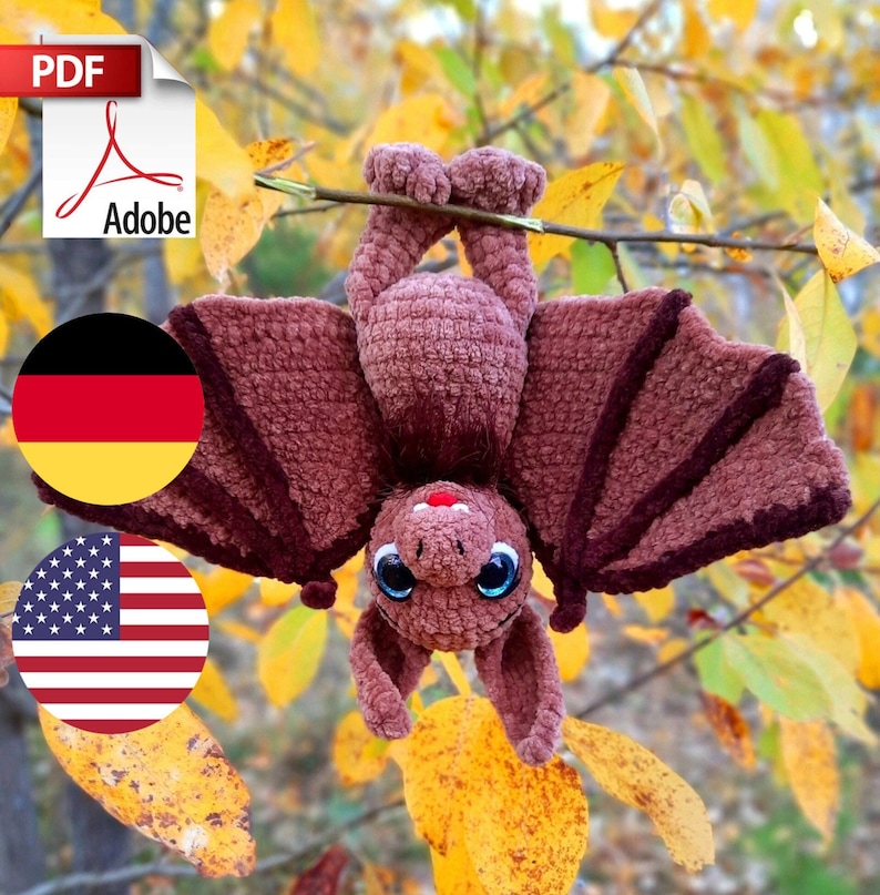 Pu&ograve; includere: Un pipistrello di uncinetto marrone appeso a un ramo con foglie autunnali. Il pipistrello ha grandi occhi blu e un naso rosso. Il pipistrello &egrave; a testa in gi&ugrave; con le ali spiegate.
