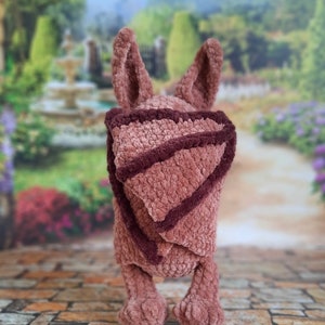 Pu&ograve; includere: Un giocattolo di peluche all'uncinetto a forma di coniglio, con orecchie lunghe e uno schema di colori rosa e marrone. Il giocattolo indossa una coperta all'uncinetto rosa e marrone.