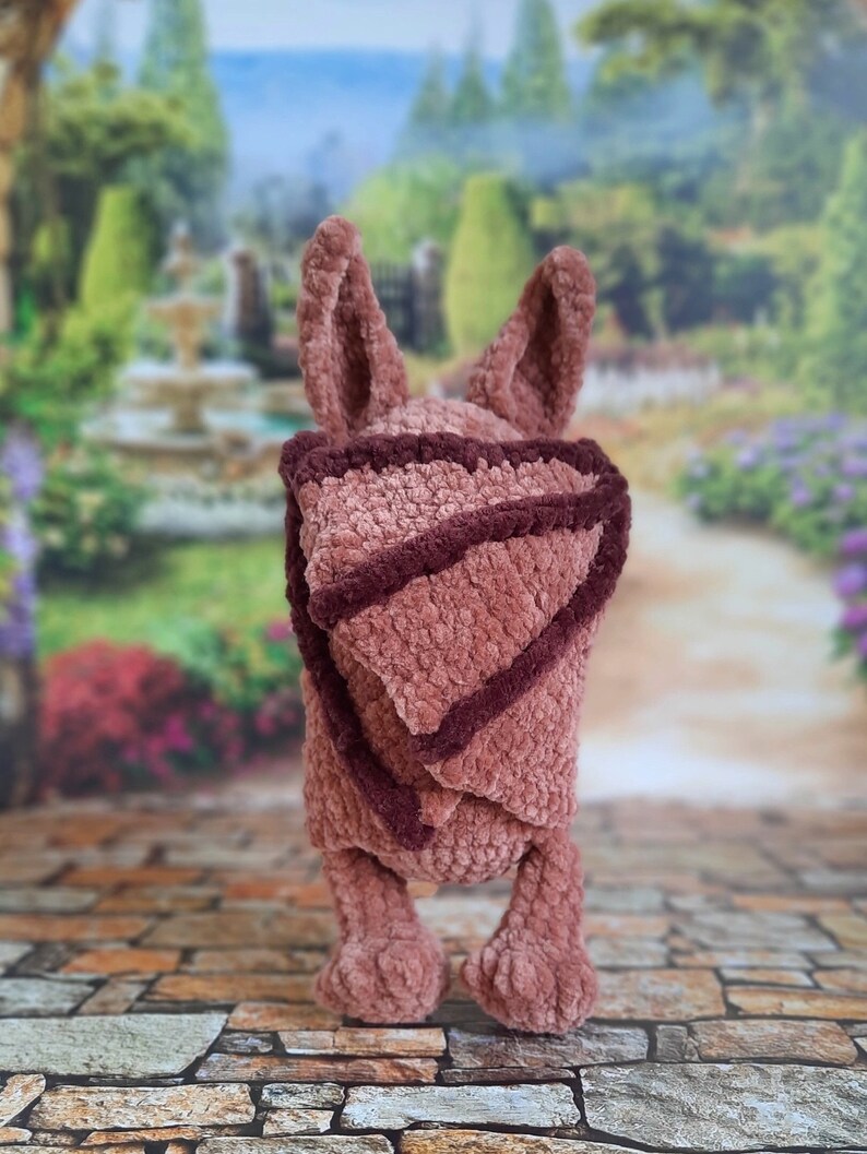 Pu&ograve; includere: Un giocattolo di peluche all'uncinetto a forma di coniglio, con orecchie lunghe e uno schema di colori rosa e marrone. Il giocattolo indossa una coperta all'uncinetto rosa e marrone.