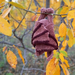 Pu&ograve; includere: Un pipistrello all'uncinetto appeso a un ramo d'albero con foglie autunnali sullo sfondo. Il pipistrello &egrave; fatto di un filato morbido e soffice in tonalit&agrave; di marrone e rosa.