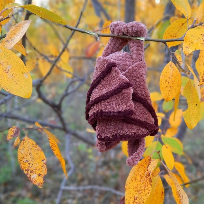 Pu&ograve; includere: Un pipistrello all'uncinetto appeso a un ramo d'albero con foglie autunnali sullo sfondo. Il pipistrello &egrave; fatto di un filato morbido e soffice in tonalit&agrave; di marrone e rosa.