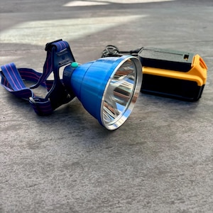 May include: A blue headlamp with a black and red strap and a silver reflector. The headlamp is connected to a yellow and black battery pack.
