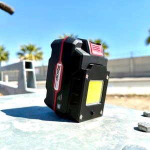 May include: Black and red X20K light with a bright yellow square light. The light has a power indicator and is sitting on a metal surface. The background is a bright blue sky with palm trees.