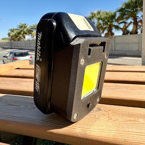 May include: A black Makita 18V 2.0Ah battery with a black 3D-printed light attachment. The light has a bright yellow square LED panel. The battery and light are resting on a wooden surface, with a street and palm trees in the background.