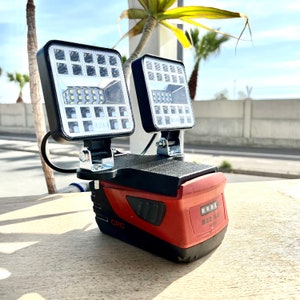 Op de afbeelding: Een rood en zwart batterij-aangedreven werklicht met twee LED-panelen. Het licht heeft een zwarte handgreep en een rode batterijpack met de tekst "B22 5.2 Li-ion" erop gedrukt.