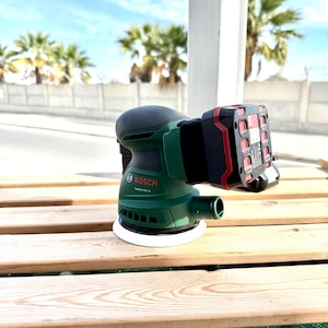 May include: A green and black Bosch random orbit sander with a red and black battery pack attached. The sander has a white sanding pad and is sitting on a wooden surface. The Bosch logo is visible on the sander.
