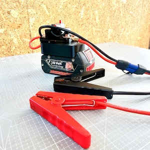 Op de afbeelding: Een zwarte en rode 18-volt batterij-startbooster met rode en zwarte kabels en klemmen. De rode klem staat op de voorgrond en de zwarte klem erachter. De startbooster staat op een grijze ondergrond.