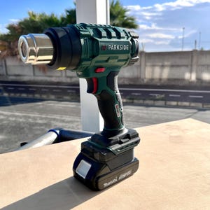 May include: A green and black Parkside heat gun with a silver nozzle. The tool has a red trigger and a black handle. A black Makita battery is attached to the base. The tool is sitting on a light brown surface.