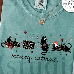 May include: A seafoam green t-shirt with a graphic of three black cats decorated with Christmas lights and a Santa hat. The text "merry catmas" is below the cats. The shirt also has the text "digital download" and "designed by kstudio".