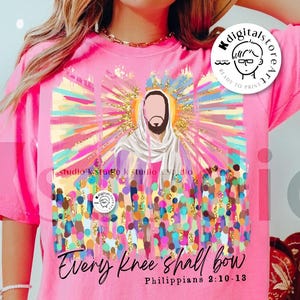May include: A pink t-shirt with a colorful graphic design featuring a person with a halo and the text "Every knee shall bow Philippians 2:10-13".