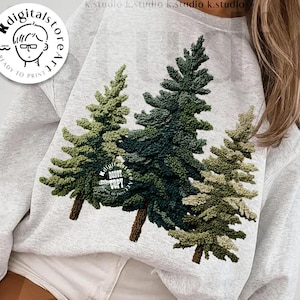 May include: A light gray sweatshirt featuring a detailed embroidered design of three evergreen trees in shades of green and brown. The words "digital download" and "designed by Kstudio" are printed on the bottom of the image. A circular logo is in the upper left corner.