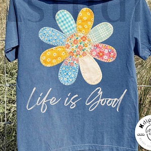 Puede incluir: Camiseta azul con una gran flor colorida aplicada. La flor tiene pétalos de varios patrones y colores, con un centro floral. Las palabras "Life is Good" están impresas debajo. La camiseta está colgada en un tendedero.