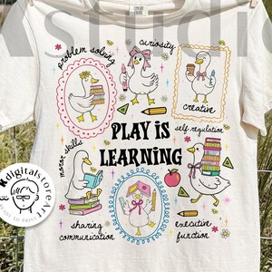 May include: Cream-colored t-shirt with a playful design featuring cartoon ducks and the text "PLAY IS LEARNING." The shirt includes illustrations of ducks with books, pencils, and an apple, promoting learning and creativity. The words "digital download" are also visible.