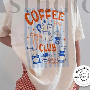 Puede incluir: Camiseta color crema con un gráfico de "COFFEE CLUB" en naranja y azul. El diseño incluye ilustraciones relacionadas con el café, como una cafetera, una taza y un molinillo. También tiene el texto "day every day".