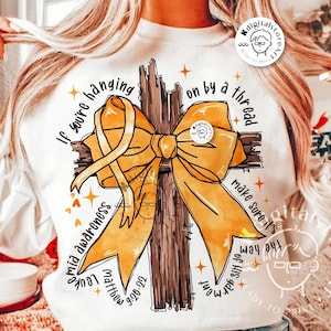 May include: A white sweatshirt with a yellow ribbon tied in a bow around a brown wooden cross. The text "If you're hanging on by a thread make sure it's the hem of His garment" is printed around the cross. The text "Matthew 9:20-22" is printed below the cross.