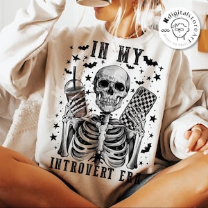 May include: A white sweatshirt with a black and white graphic of a skeleton holding a phone and a cup. The text "IN MY INTROVERT ERA" is printed on the sweatshirt.
