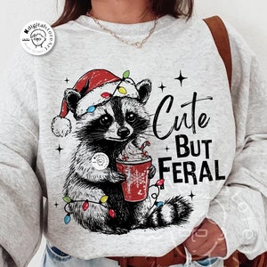 Può includere: Una felpa grigia con un adorabile procione che indossa un cappello da Babbo Natale e tiene una tazza di cioccolata calda. Il procione è circondato da luci natalizie. Il testo "Cute But Feral" è stampato sulla felpa.
