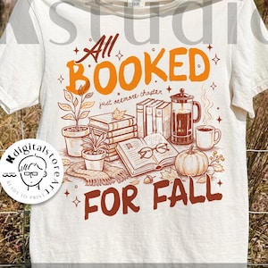 May include: Cream-colored t-shirt with the text "All Booked For Fall" in orange and brown. The design includes illustrations of books, a coffee pot, a pumpkin, and plants, evoking an autumn theme. The shirt is hanging on a clothesline.