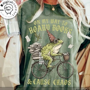 May include: Green t-shirt with a whimsical illustration of a frog riding a bicycle, wearing a wizard hat, and carrying books. Text reads "ON MY WAY TO HOARD BOOKS & CAUSE CHAOS." The design includes a dragon and flying papers. "Digital Download" is printed at the bottom.
