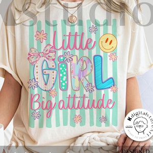May include: A cream-coloured t-shirt featuring a graphic print with vertical teal stripes, a pink bow, a yellow smiley face, and the text Little Girl Big attitude in pink and blue. The design includes various small flower illustrations.