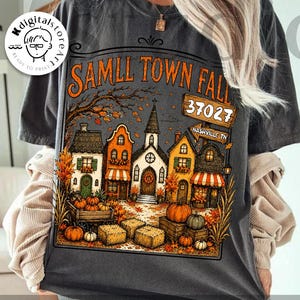 May include: A charcoal grey t-shirt featuring a graphic print of a small town during autumn. The design includes houses, pumpkins, hay bales, and a sign that reads SAMLL TOWN FALL 37027 NASHVILLE TN. The illustration uses orange, yellow, and brown tones.