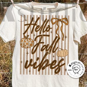 Puede incluir: Camiseta de color crema con un diseño temático de otoño. El diseño incluye el texto "Hello Fall Vibes" en escritura marrón, junto con calabazas y un lazo. La camiseta está colgada en un tendedero.