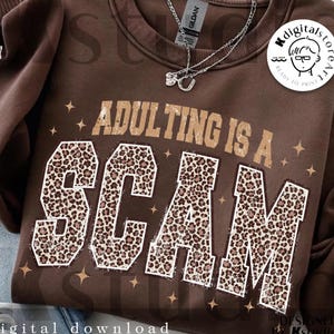 May include: A brown sweatshirt with the phrase "ADULTING IS A SCAM" in a tan, distressed font. The letters of "SCAM" are filled with a leopard print pattern. A silver necklace is draped across the top.