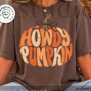 May include: A dark brown short-sleeve t-shirt featuring a large graphic of an orange pumpkin with a brown stem and a small bow. The text "HOWDY PUMPKIN" is printed in a distressed, bold, rounded font across the center of the pumpkin illustration.