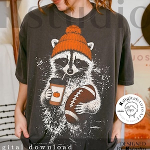 May include: Dark gray t-shirt featuring a raccoon wearing an orange beanie and holding a football and a pumpkin spice latte. The shirt has a distressed, vintage look with the words "digital download" at the bottom.