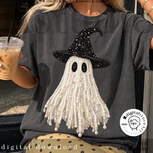 May include: A charcoal gray t-shirt featuring a central graphic of a ghost wearing a witch hat. The ghost is embellished with white pearls and clear beads, while the hat is decorated with black beads. The design has a textured, beaded appearance.