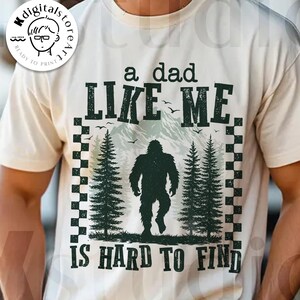 Pode incluir: Camiseta creme com um gráfico que apresenta um Bigfoot, árvores e montanhas. O texto diz "A Dad Like Me Is Hard To Find". O design é emoldurado com um padrão xadrez. "Digital Download" é impresso na parte inferior.