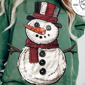 May include: Green sweatshirt featuring a stitched snowman design. The snowman has a top hat, carrot nose, and a red scarf. The design includes the text "DIGITAL DOWNLOAD" and "DESIGNED BY Kstudio".