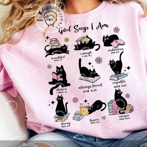 Puede incluir: Sudadera rosa con el texto "God Says I Am" y un diseño de gatos negros con libros y versículos bíblicos.