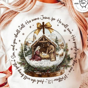 May include: White sweatshirt with a snow globe design featuring a nativity scene. The text around the globe reads "And you shall call His name Jesus, for He will save His people from their sins - Matthew 1:21".