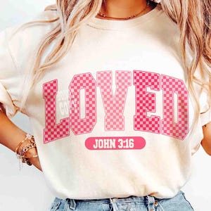 May include: Cream-colored sweatshirt with the word "LOVED" in pink, checkered letters. Below the word is the text "JOHN 3:16" in a pink rectangle. The sweatshirt has long sleeves.