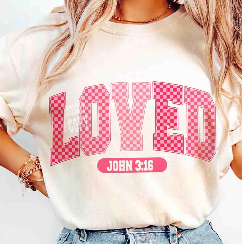 May include: Cream-colored sweatshirt with the word "LOVED" in pink, checkered letters. Below the word is the text "JOHN 3:16" in a pink rectangle. The sweatshirt has long sleeves.