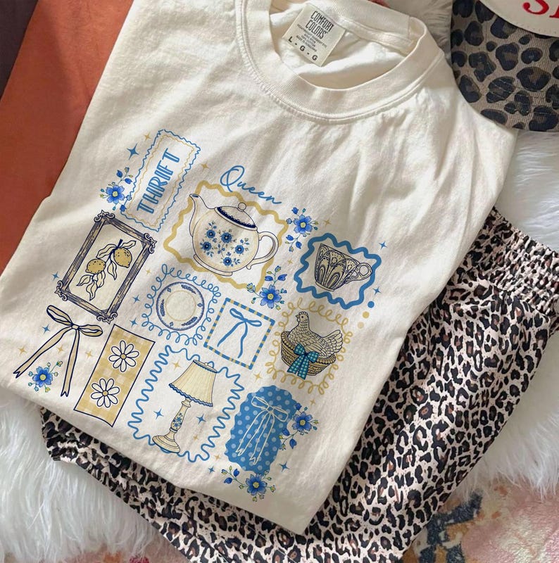 May include: Cream-colored t-shirt with a collage of blue and yellow illustrations, including a teapot, lemons, and bows. The word "Thrift" is visible. Paired with leopard print pants, the shirt has a relaxed, vintage aesthetic.